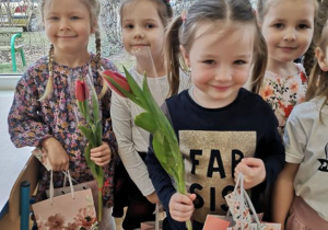 Widok na cztery dziewczynki pozujace do zdjęcia z kwiatami i prezentami.